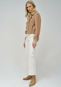 Chaqueta beige corta, de doble botonadura, con cuello con muescas y cinturón. Combinada con pantalones blancos de cintura alta y botines beige con punta afilada.