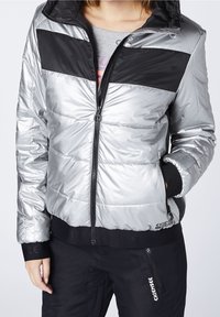 Silverfärgad puffjacka med svarta horisontella ränder, dragkedja fram och ribbade muddslut. Har en slät yta och sidofickor.