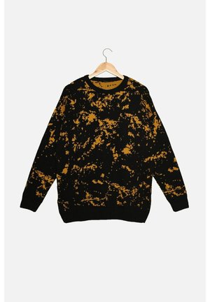 Pull en tricot noir avec un motif éclaboussé irrégulier jaune moutarde, manches longues, col rond, suspendu à un cintre en bois sur fond blanc.