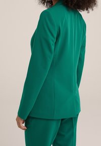 Zelený dokonale padnoucí blejzr se strukturovaným střihem, zapínáním na jeden knoflík a hladkým, texturovaným materiálem, doplněný o ladící kalhoty.