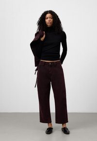 Donna con capelli ricci che indossa un dolcevita nero e pantaloni larghi bordeaux scuri, tiene una giacca coordinata sulla spalla, in piedi contro un muro semplice.