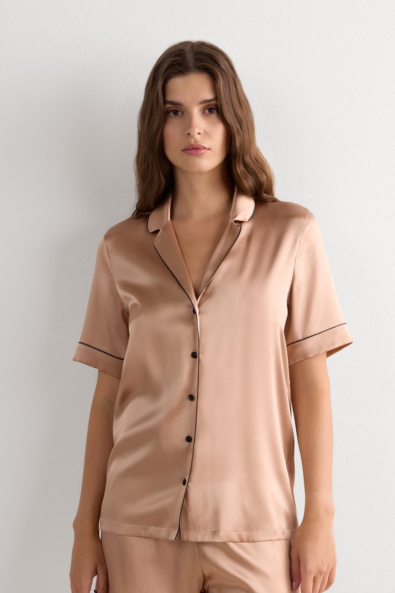 Vrouw met lang bruin haar draagt een lichtbruine satijnen korte mouwen pyjamabovenstuk met zwarte knopen en zwarte bies, staand tegen een witte muur.
