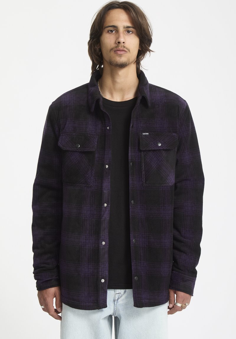 Camisa de franela a cuadros en negro y morado con botones en la parte delantera, dos bolsillos en el pecho y una textura suave. Llevada sobre una camiseta negra.