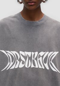 Felpa grigia con un design grafico testuale bianco ondulato. Collo a giro con tessuto testurizzato e dettagli superficiali sottili.