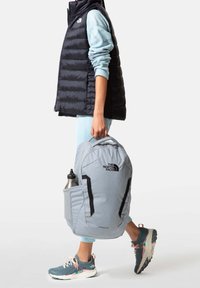 Grau farbener Rucksack mit schwarzen Akzenten, ausgestattet mit einer Seitentasche für eine Wasserflasche und einem Logo auf der Vorderseite. Wird von einer Person in Sportbekleidung getragen.