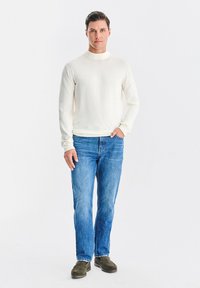 Kremowy top z długim rękawem i stójką, zestawiony z niebieskimi dżinsami oraz oliwkowozielonymi butami wsuwanymi. Gładka faktura, luźny fason.