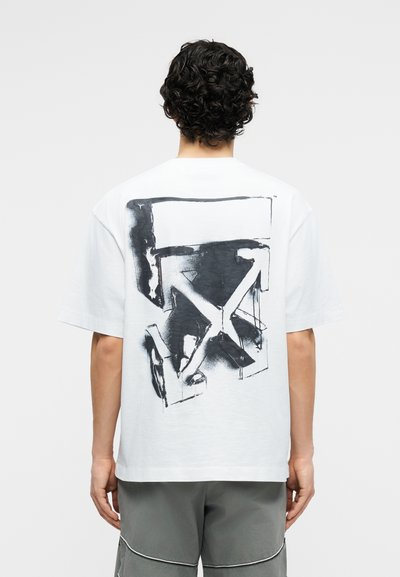 Άνδρας με σκούρα, σγουρά μαλλιά, που φοράει λευκό, φαρδύ T-shirt με μαύρο αφηρημένο γραφικό βέλος στην πλάτη και γκρι σορτς, στέκεται με την πλάτη στραμμένη.