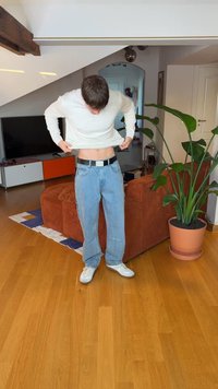 Oseba v svetli majici z dolgimi rokavi, širokih modrih kavbojkah in belih supergah, ki si prilagaja majico, v ozadju je lončnica.