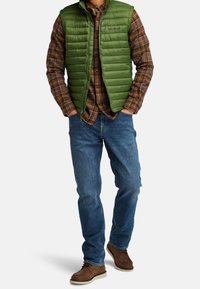 Grön vadderad väst över en brun rutiga skjorta, blå jeans och bruna snörskor. Väst har dragkedja och synlig logotyp.