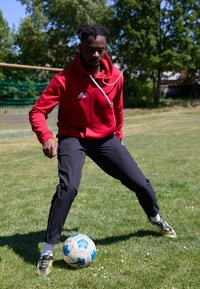 Roter Pullover mit weißen Akzenten und einem Adidas-Logo, schwarze Sporthosen und schwarze Fußballschuhe kontrollieren einen blau-weißen Fußball auf Gras.