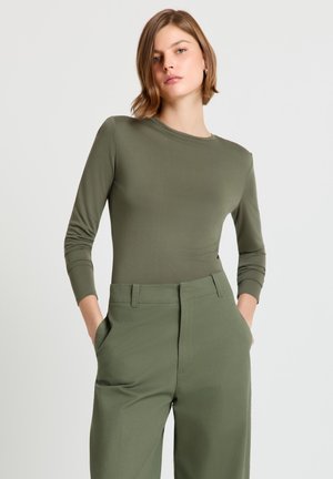 Femme aux cheveux courts châtain clair portant un haut manches longues vert olive et un pantalon taille haute assorti, mains dans les poches, arrière-plan neutre.
