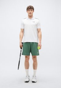 Hvit polotrøye med svarte sidestriper, grønne shorts, holder en tenniskølle og en gul tennisball, med hvite sokker og sko.
