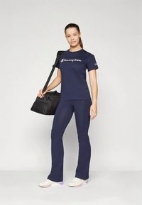 Vrouw in een marineblauw Champion T-shirt en broek, met een zwarte sporttas over haar schouder, staand tegen een effen achtergrond.