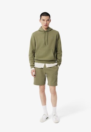 Hoodie vert olive avec une poche kangourou, associé à un short assorti. Il est accompagné d'un t-shirt à rayures blanches en dessous et de baskets blanches.
