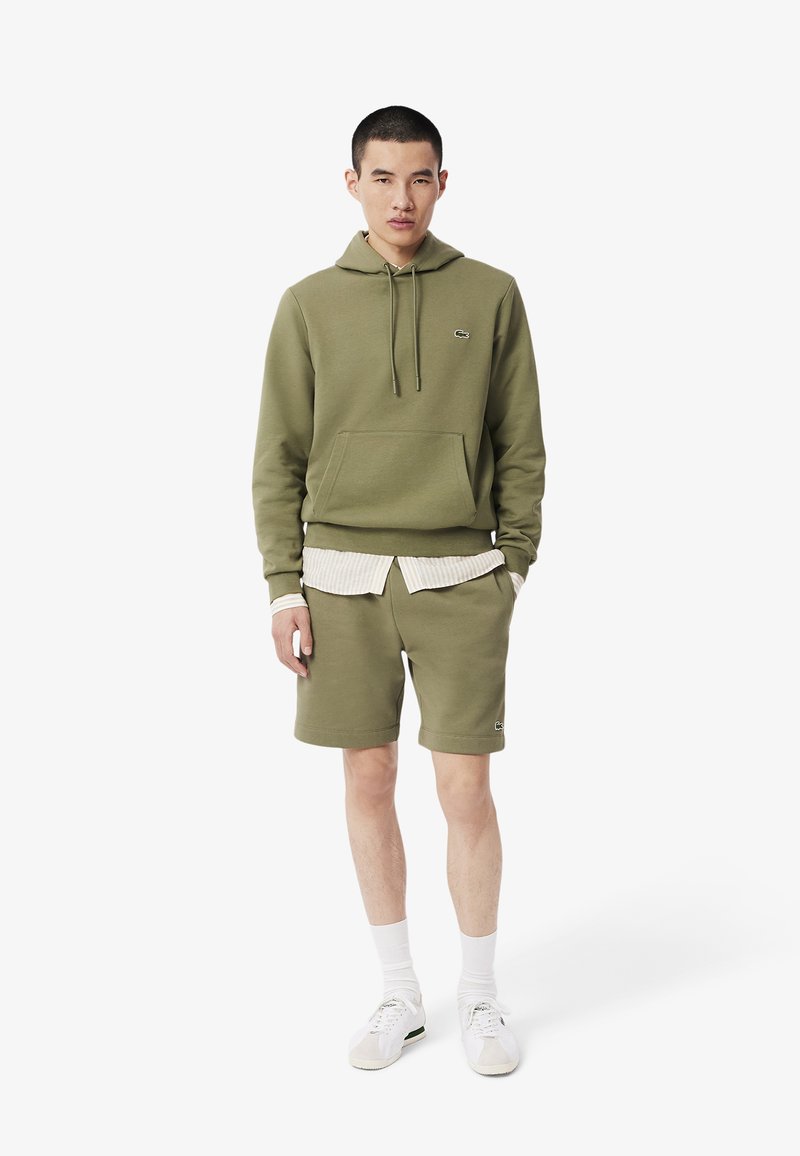 Sudadera color verde oliva con un bolsillo tipo canguro, combinada con pantalones cortos a juego. Presenta una camiseta de rayas blancas debajo y zapatillas blancas.
