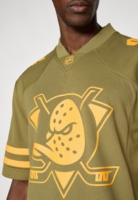 Olivově zelený hokejový dres se velkým oranžovým grafickým designem hlavy kachny, s žlutými pruhy na rukávech a logem NHL na límci.