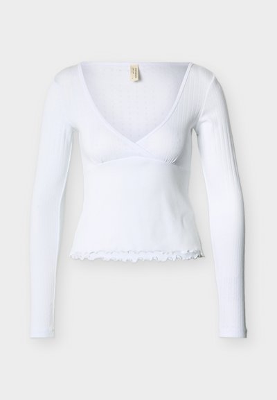 Λευκό ριμπ τοπ με μακριά μανίκια, βαθύ V λαιμόκοψη, τύπου περιτύλιξης στο μπροστινό μέρος και τελείωμα με κυματιστή άκρη σε μονόχρωμο φόντο.