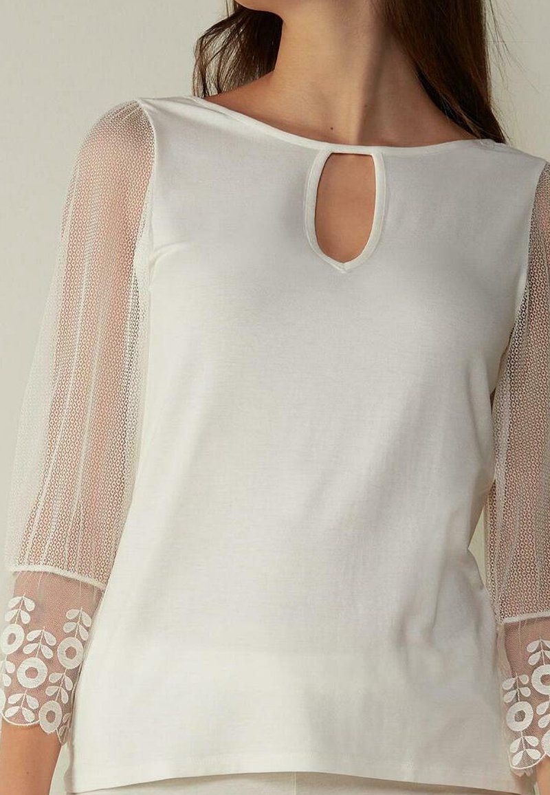 Top bianco con scollatura a forma di chiave, caratterizzato da maniche in pizzo trasparente decorate con ricami floreali sui polsini. Texture liscia e vestibilità comoda.