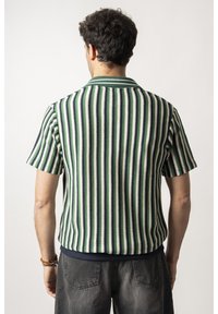 La chemise à manches courtes présente des rayures verticales vertes, bleu marine et blanches, avec un col côtelé et un ourlet bleu marine uni. Le tissu semble texturé.