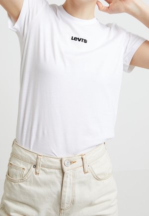 Personne portant un t-shirt blanc Levi's rentré dans un pantalon écru, levant les bras avec les mains près de la tête sur un fond uni.