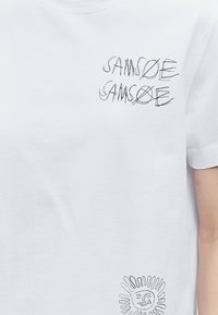 Λευκό T-shirt με μαύρο κείμενο σε στυλ σκίτσου "SAMSOE" επαναλαμβανόμενο δύο φορές και μια απεικόνιση ήλιου κοντά στο κάτω δεξιό μέρος.