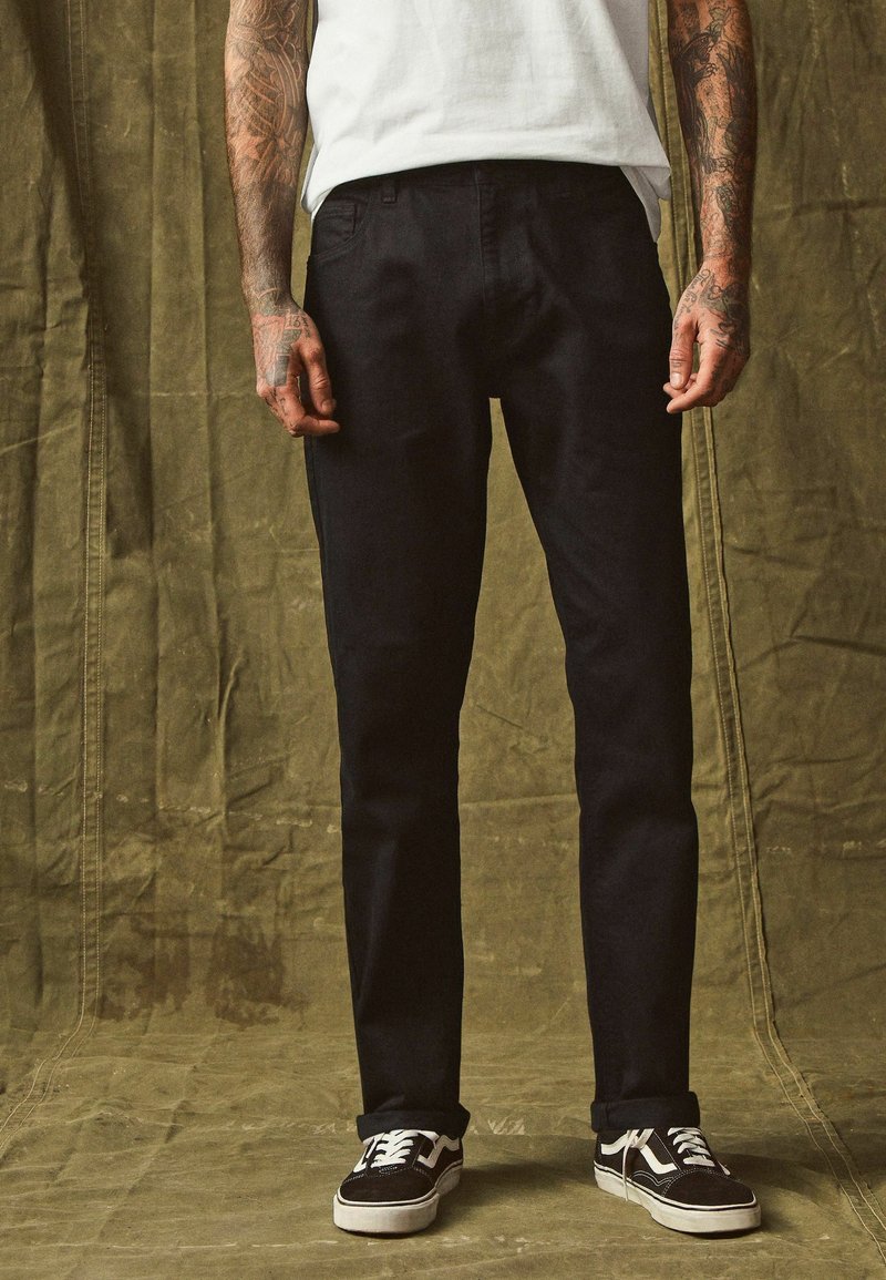 Zwarte denim broek met een recht model, voorzien van vijf zakken, gedragen met zwart-witte sneakers. De textuur lijkt glad en stevig.