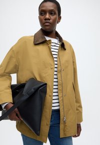 Veste kaki avec col marron, fermeture éclair à l'avant et boutons-pression ; sac fourre-tout noir ; chemise rayée ; jean bleu. Design simple et fonctionnel.