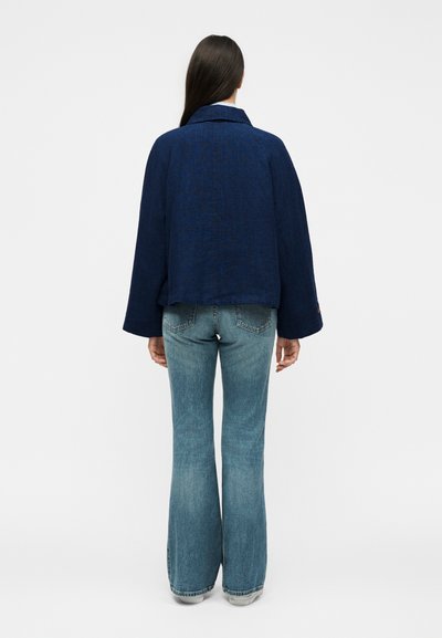 Γυναίκα με μακριά σκούρα μαλλιά που φοράει σκούρο μπλε μπουφάν και ανοιχτό μπλε καμπάνα τζιν, όρθια με την πλάτη προς ένα απλό λευκό φόντο.