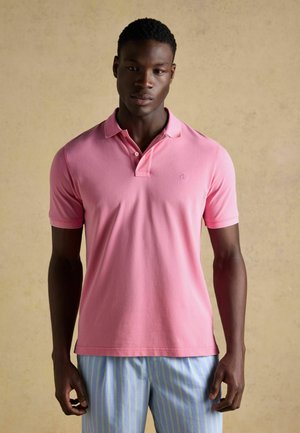 Junger Mann trägt ein rosa Poloshirt und hellblaue Hosen mit gelben Streifen, steht vor einem einfarbigen beigen Hintergrund.