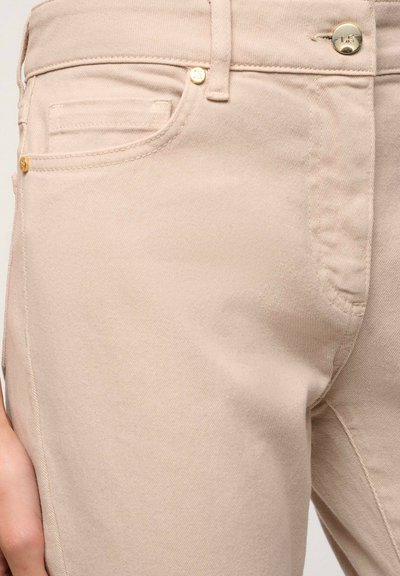 Beige puuvillahousut, joissa on sileä rakenne, etutasku, kultasävyisiä yksityiskohtia ja klassinen viisi taskua -design.