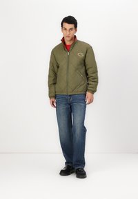 Quiltet olivengrøn jakke med høj krave, lynlås lukning og logo på brystet, parret med blå jeans og sorte sko.