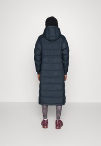 Lang, mørk puffjakke med hette, med quilte-seksjoner. Brukt over mønstrede leggings og brune sneakers med glidelåser på sidene.