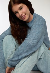 Jasnoniebieski sweter robiony na drutach z wysokim kołnierzem, zestawiony z jasnoniebieskimi dżinsami. Sweter ma teksturowany splot. Ogólny wygląd casualowy.