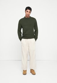 Donkergroen gebreid sweater met geribbelde manchetten en zoom, gecombineerd met loszittende crème broek en tan schoenen. Effen achtergrond.