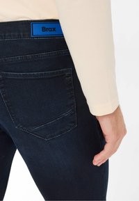 Donkerblauwe denim jeans met een zichtbare achterzak. Brax-logo in het blauw op de tailleband. De stof lijkt glad met een lichte textuur.