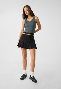 Žena v šedém bezrukávovém crop topu, černé plisované mini sukni s páskem v pase, bílých ponožkách a černých mokasínech na jednobarevném pozadí.
