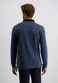Mann mit kurzen braunen Haaren, der ein blaues Langarmpoloshirt und dunkle Hosen trägt, von hinten vor einem schlichten Hintergrund gezeigt.