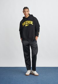 Fekete kapucnis pulóver sárga "Reebok" logóval, fekete nadrággal és fehér sportcipővel párosítva. Simább anyag, laza szabás, és elülső zseb a kapucnis pulóveren.