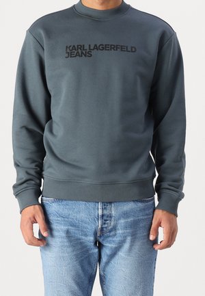 Mężczyzna w ciemnoszarej bluzie z napisem „KARL LAGERFELD JEANS” i jasno-niebieskich dżinsach, stojący na jednolitym tle.