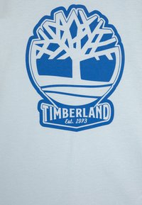 Bijela pamučna majica s velikim plavim Timberland logotipom i grafikom drveta te natpisom "Osnovano 1973" u podebljanom fontu.