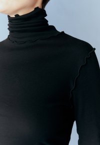 Personne portant un col roulé côtelé noir avec des bords froncés au niveau du col et de l'épaule sur un fond gris clair.