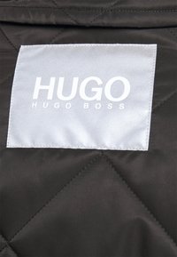 Tissu matelassé noir avec une étiquette rectangulaire blanche cousue affichant "HUGO HUGO BOSS" en lettres blanches en gras.