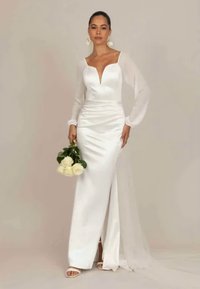 Vestido longo de cetim branco com decote profundo, mangas bufantes transparentes e fenda lateral. A modelo segura um bouquet de rosas brancas.