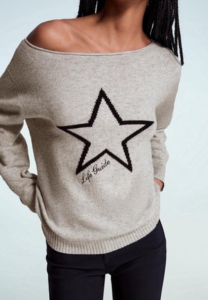 Beżowy sweter z odkrytymi ramionami z dużą czarną gwiazdą i napisem „Life Guide”, noszony z czarnymi spodniami, widoczny od szyi do bioder.