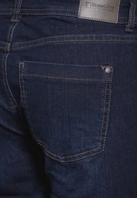 Mörkblå denimjeans med en bakficka som har en silvernagl. Sömmarna är i en kontrasterande ljusare färg. Läderetikett vid midjan.