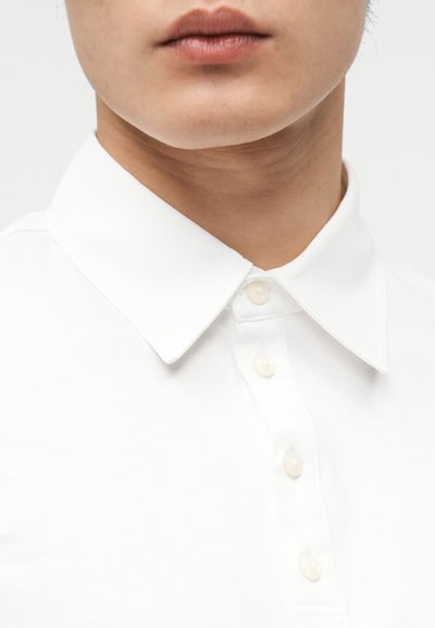 Κοντινό πλάνο ενός ατόμου που φοράει λευκό πουκάμισο με γιακά κουμπωμένο μέχρι επάνω, εμφανίζοντας τα χείλη, το πηγούνι και τον λαιμό πάνω σε απλό φόντο.