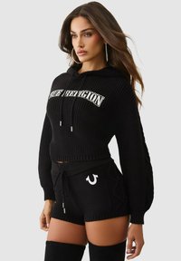 Schwarzer, gestrickter, kurz geschnittener Hoodie mit Logo, kombiniert mit schwarzen Shorts, die ein seitliches Design und einen Kordelzug haben. Dazu passend schwarze Overknee-Stiefel.
