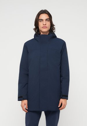 Young person with shoulder-length dark hair wearing a dark blue hooded winter coat standing straight, facing forward.