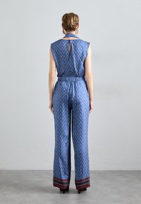 Modra vzorirana kombinezonska obleka s ovratnikom in brezrokavnim dizajnom, ki ima elastičen pas in široke hlače. Kontrastni maroon odtenki na robu.