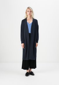 Pikk mereväe värvi kardigan avatud esisegmendiga ja kahe eesmise taskuga, seljas helesinise V-kaelusega topi ja mustade laiade pükstega. Mustad jalanõud.